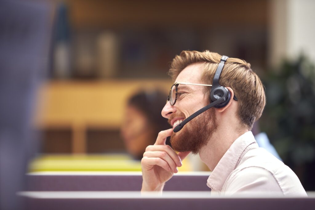 Businessman In Cubicle Wearing Headset Talking To Caller In Busy Customer Services Centre